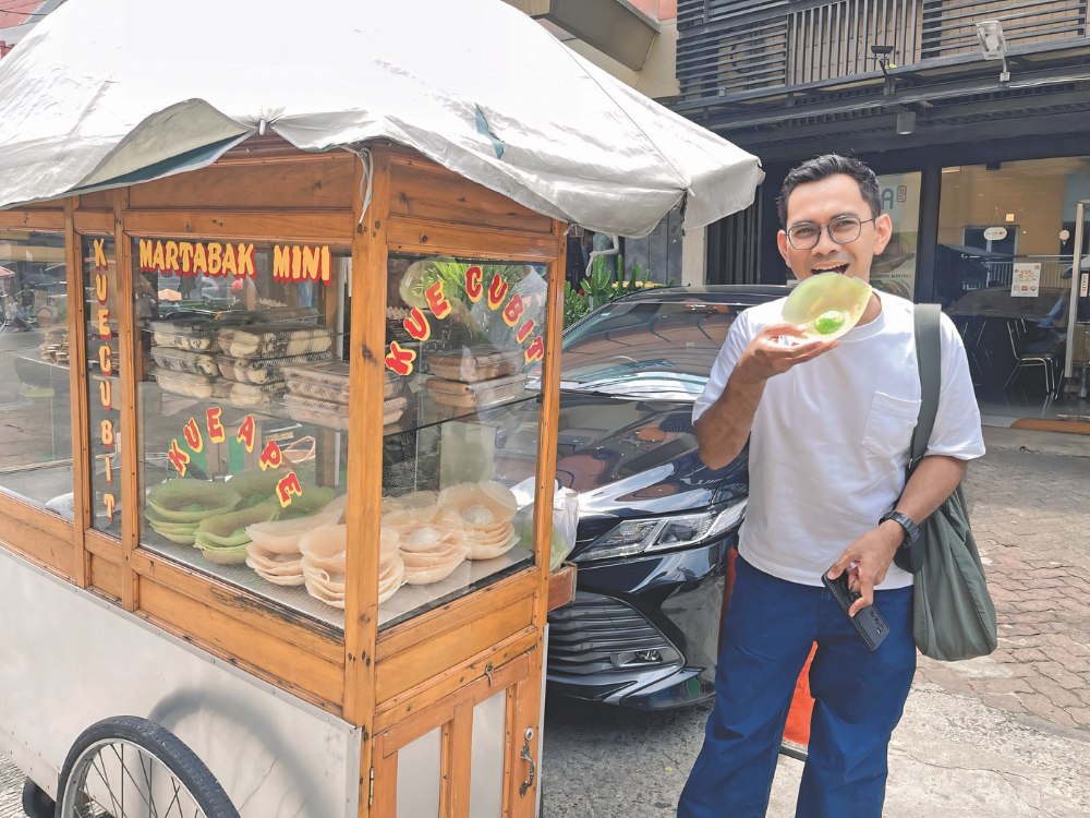 A man stands beside a street food cart labeled with "Martabak Mini" and "Kue Cubit," smiling and holding a green snack. The scene is bright and casual.