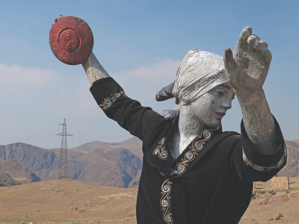 A statue of a person with silver skin, wearing a headscarf and a black, ornate garment, poised to throw a disk. Set against a mountainous landscape with a transmission tower.