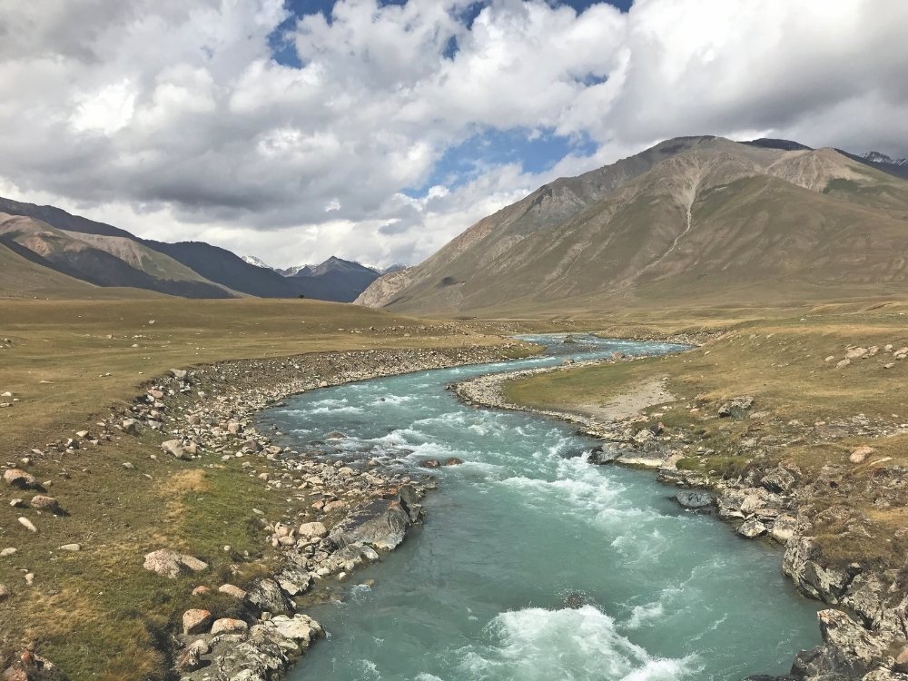 A clear blue river meanders through a vast, rocky valley under a cloudy sky, surrounded by rolling green hills and distant mountains, evoking tranquility.