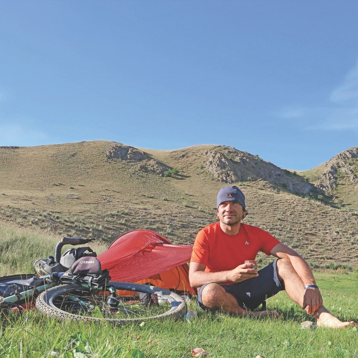 A man in a red shirt and beanie sits on grass near a red tent and bicycle. The scene is set against rolling hills and a clear blue sky, conveying a sense of adventure.