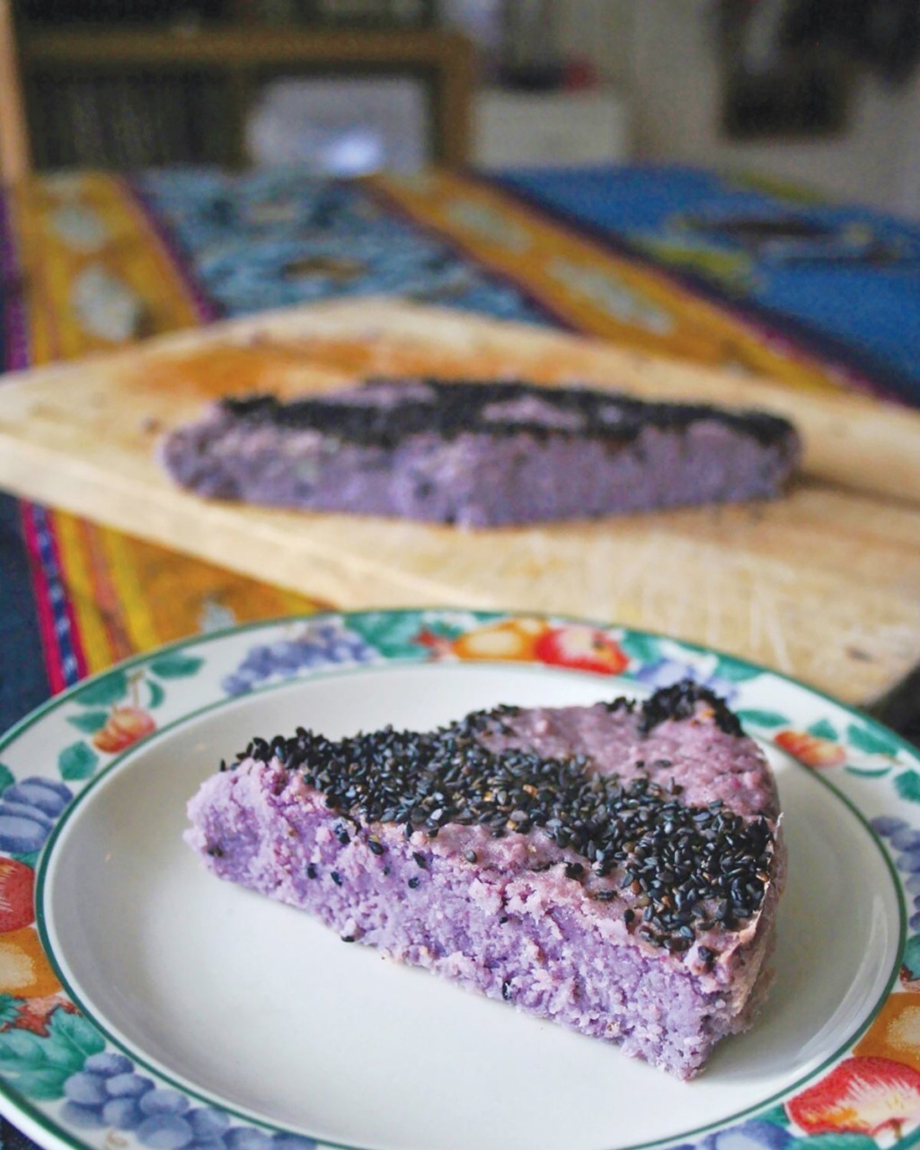 A slice of vibrant purple yam cake topped with black sesame seeds on a floral-patterned plate. Another cake slice rests on a wooden board in the background.