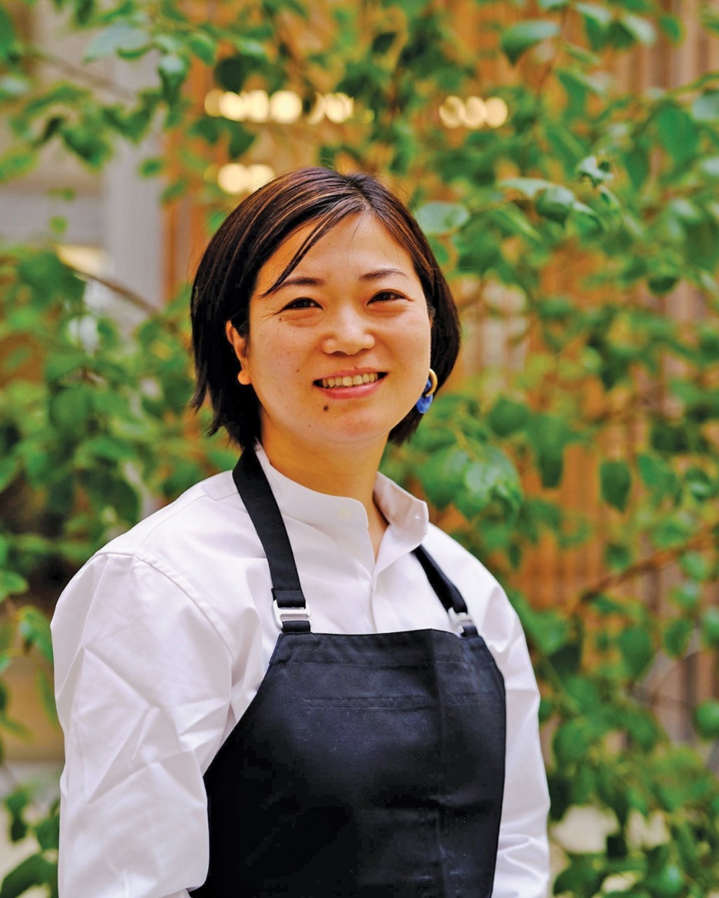 Smiling woman in a white shirt and black apron stands against a backdrop of lush green foliage. The atmosphere is bright and cheerful.