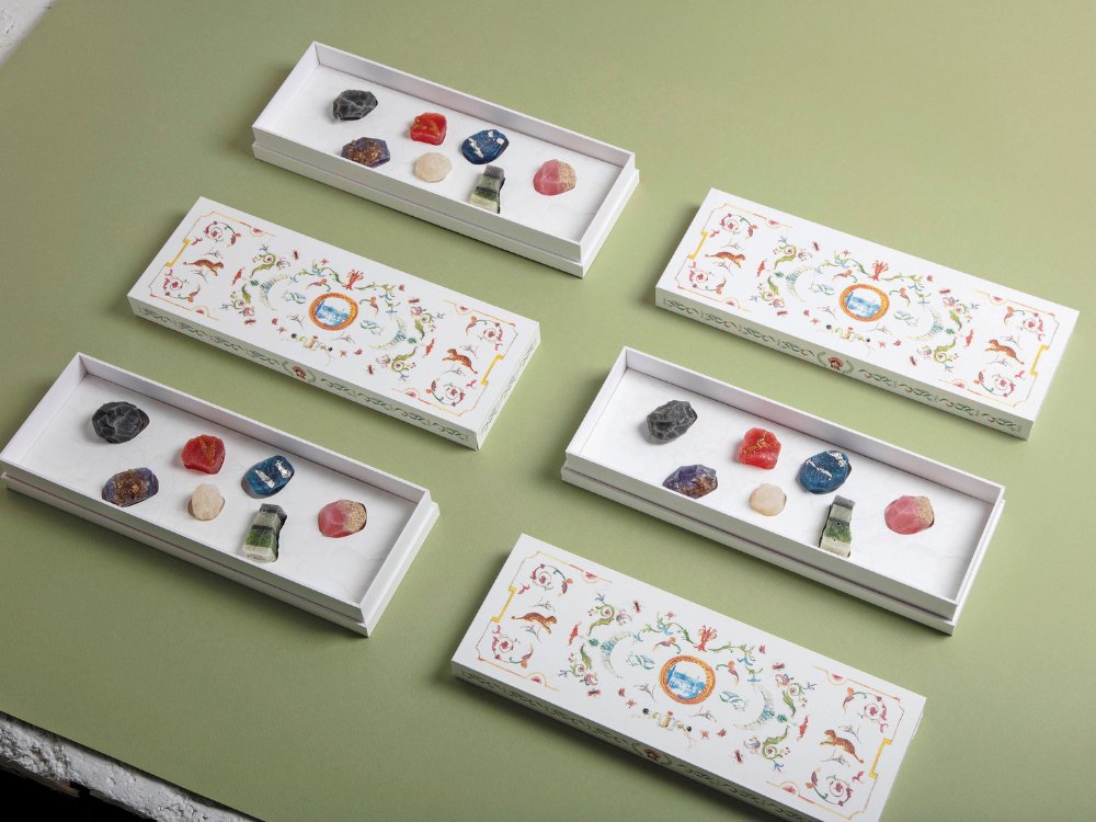 Five ornate white boxes on a green surface hold colorful gemstones, including red, blue, pink, and striped stones. The scene conveys elegance and artistry.
