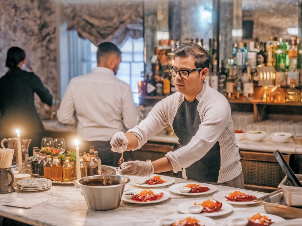 A chef in a white shirt and apron carefully garnishes plates with sauce in a stylish restaurant kitchen. Bottles and glasses adorn the background bar.