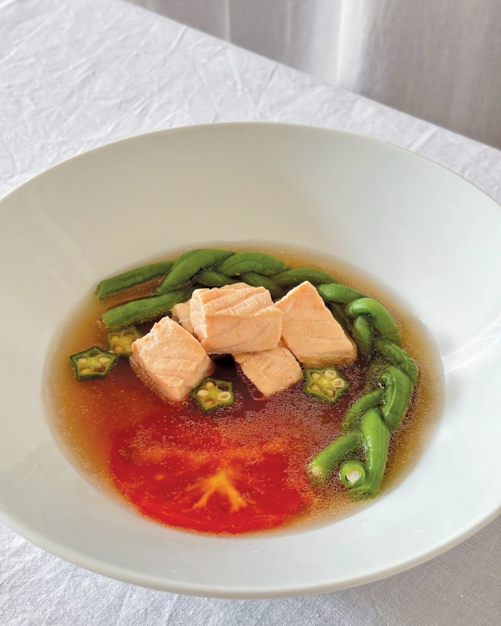 A white bowl with a broth featuring salmon cubes, okra slices, and green beans. A tomato adds a pop of red. The setting is minimalist and elegant.