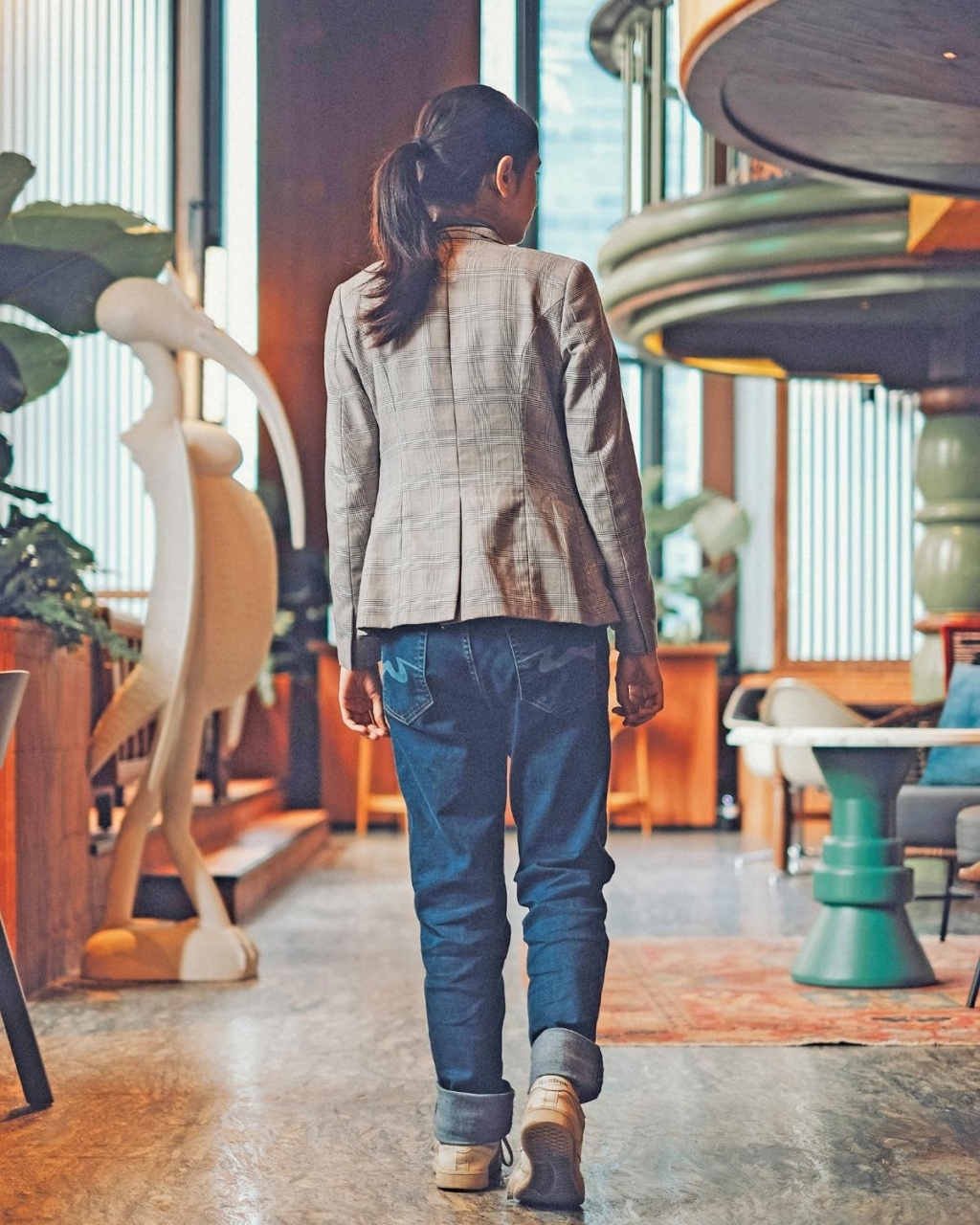 A woman with a ponytail wearing a plaid jacket and jeans walks in a stylish cafe with plants and modern decor.