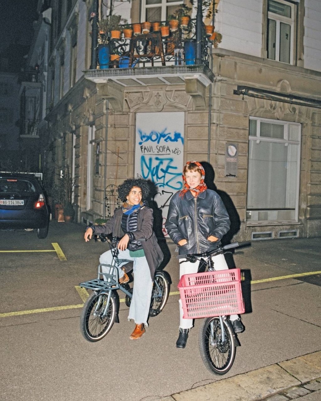 Two people pose cheerfully with bikes on a nighttime street. Graffiti, potted plants, and an iron balcony create a lively urban backdrop.