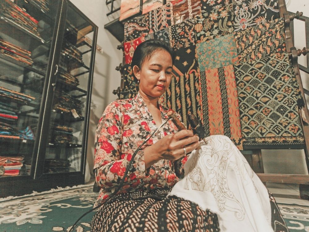 A woman in a floral blouse creates batik patterns on fabric, surrounded by vibrant textiles. The scene captures culture and artistry in a focused, serene atmosphere.