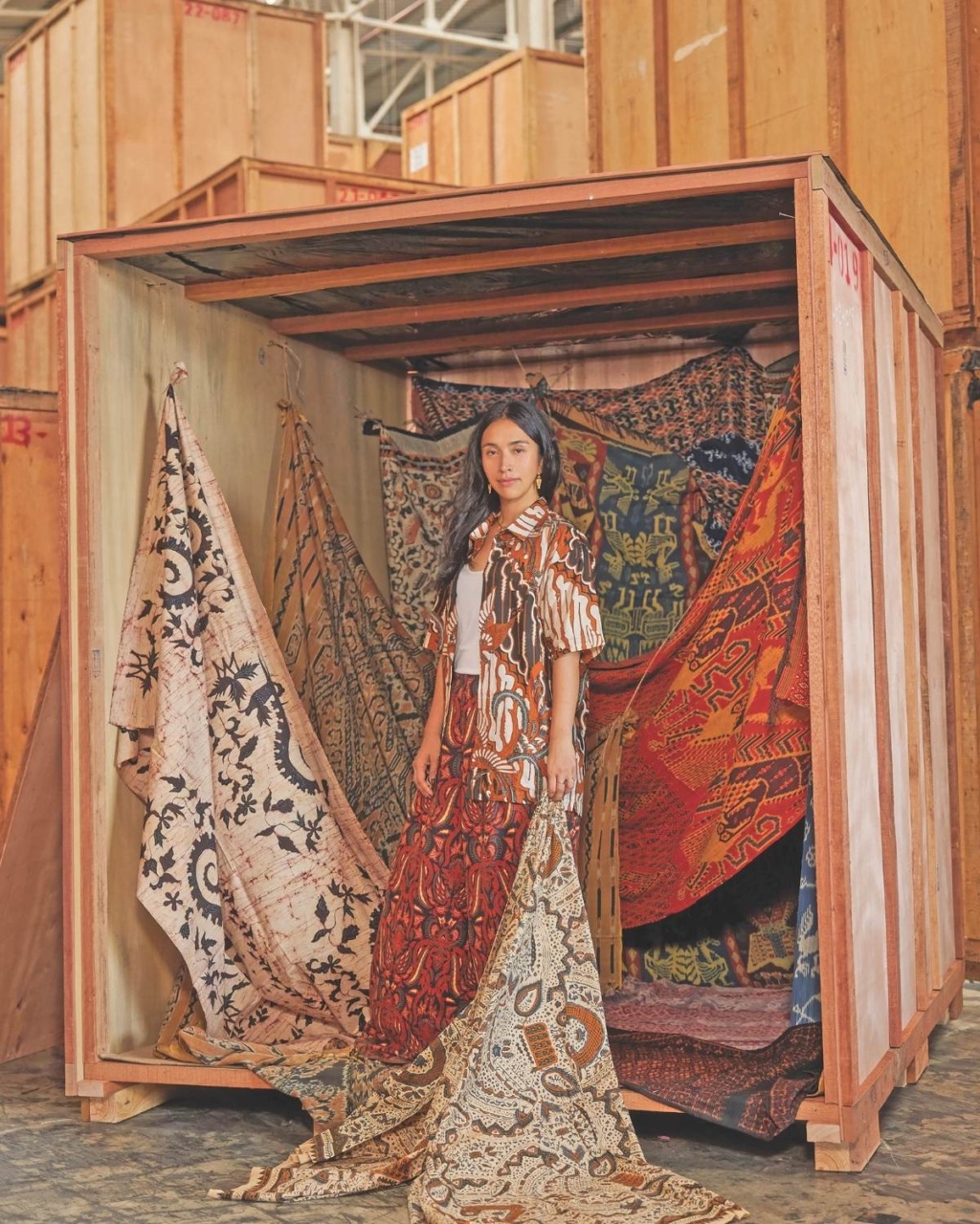 A woman stands confidently in a wooden crate surrounded by vibrant, patterned fabrics draping around her. The setting conveys an artistic, eclectic vibe.
