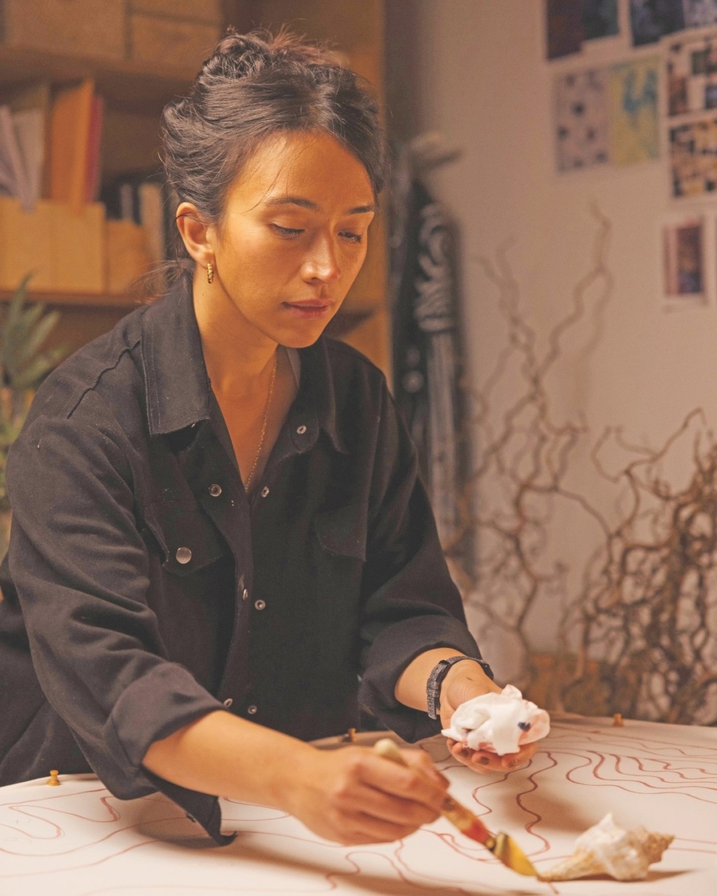 A woman in a black shirt is focused on painting wavy red lines on a canvas. The setting includes shelves with folders and artistic tools, conveying a creative atmosphere.