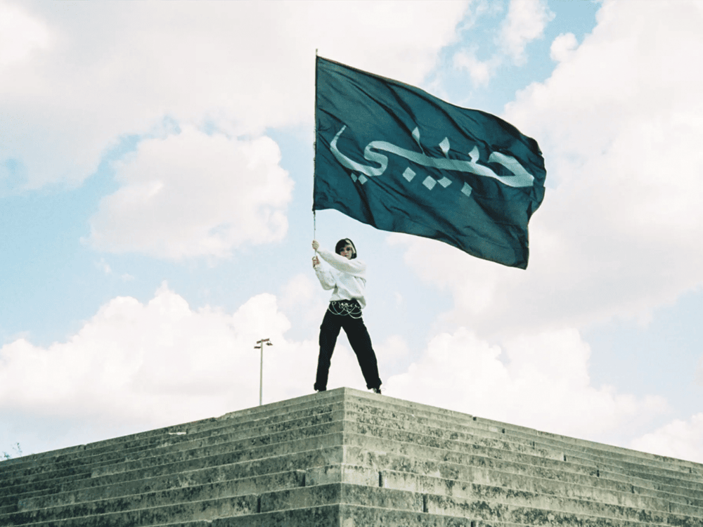 Person standing on stone steps, holding a large blue flag with white Arabic text.