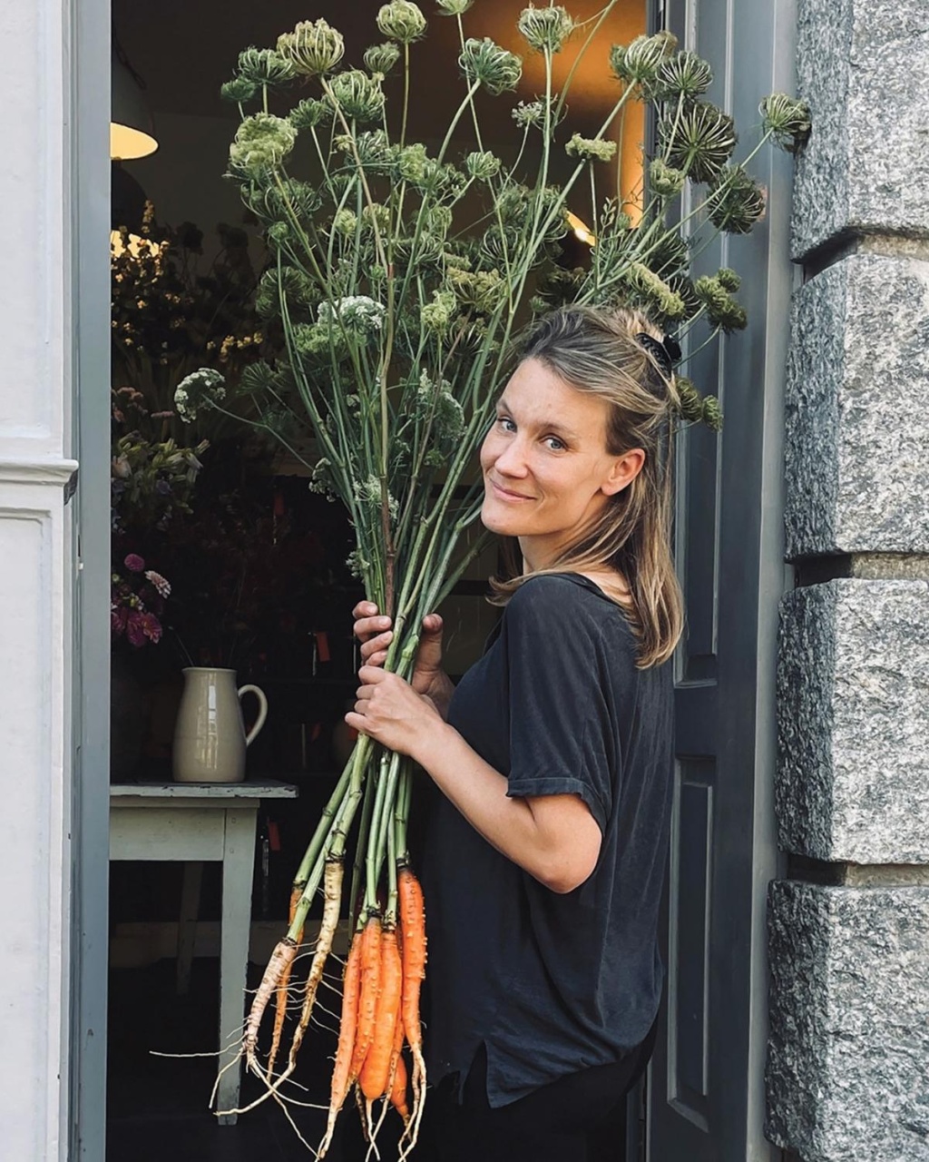 A woman smiles warmly while holding large carrots with green tops by the entrance of a building. Inside, a vase is visible on a table.