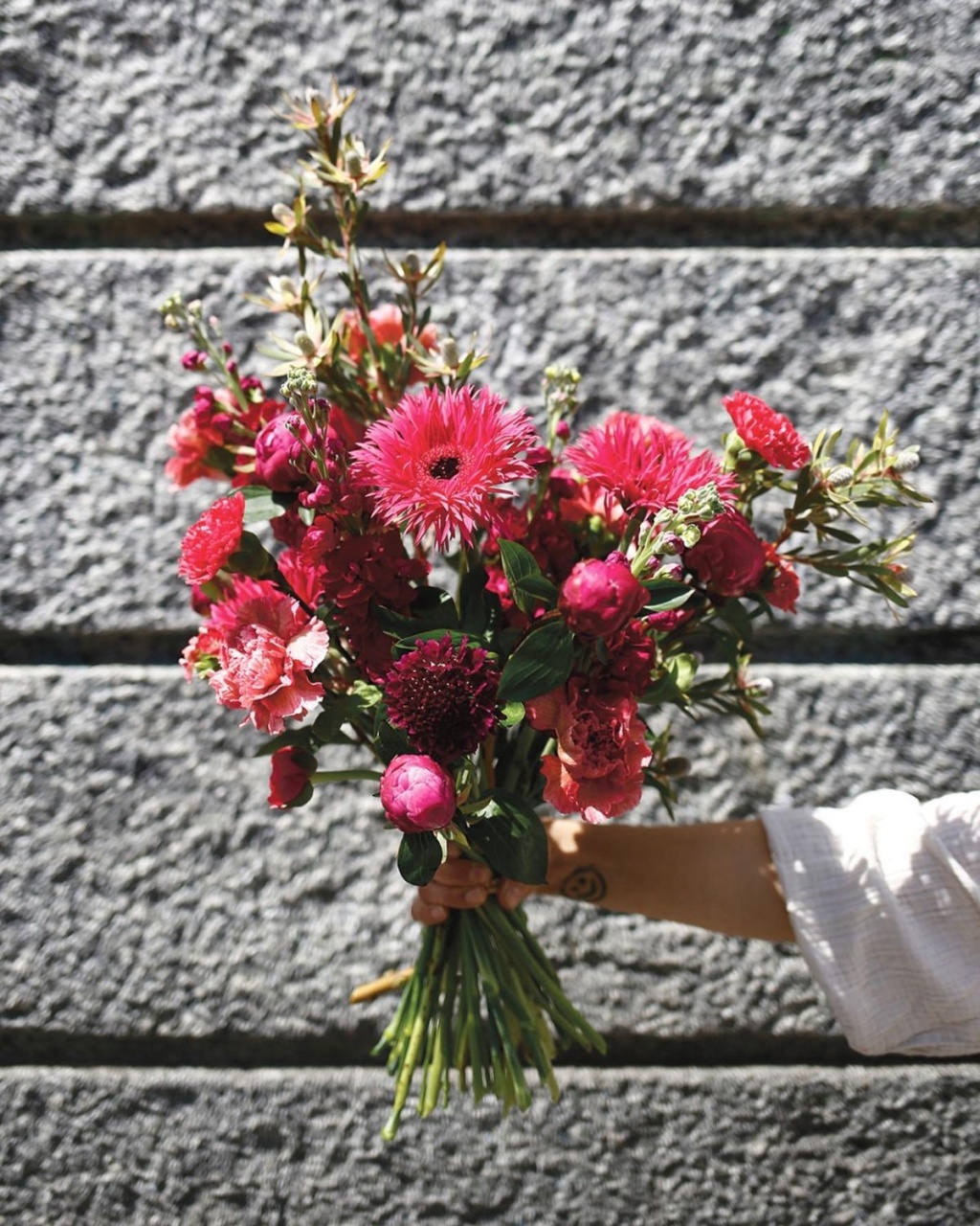 A hand holds a vibrant bouquet of pink and red flowers against a textured gray stone wall. The mix of blooms creates an uplifting, cheerful mood.