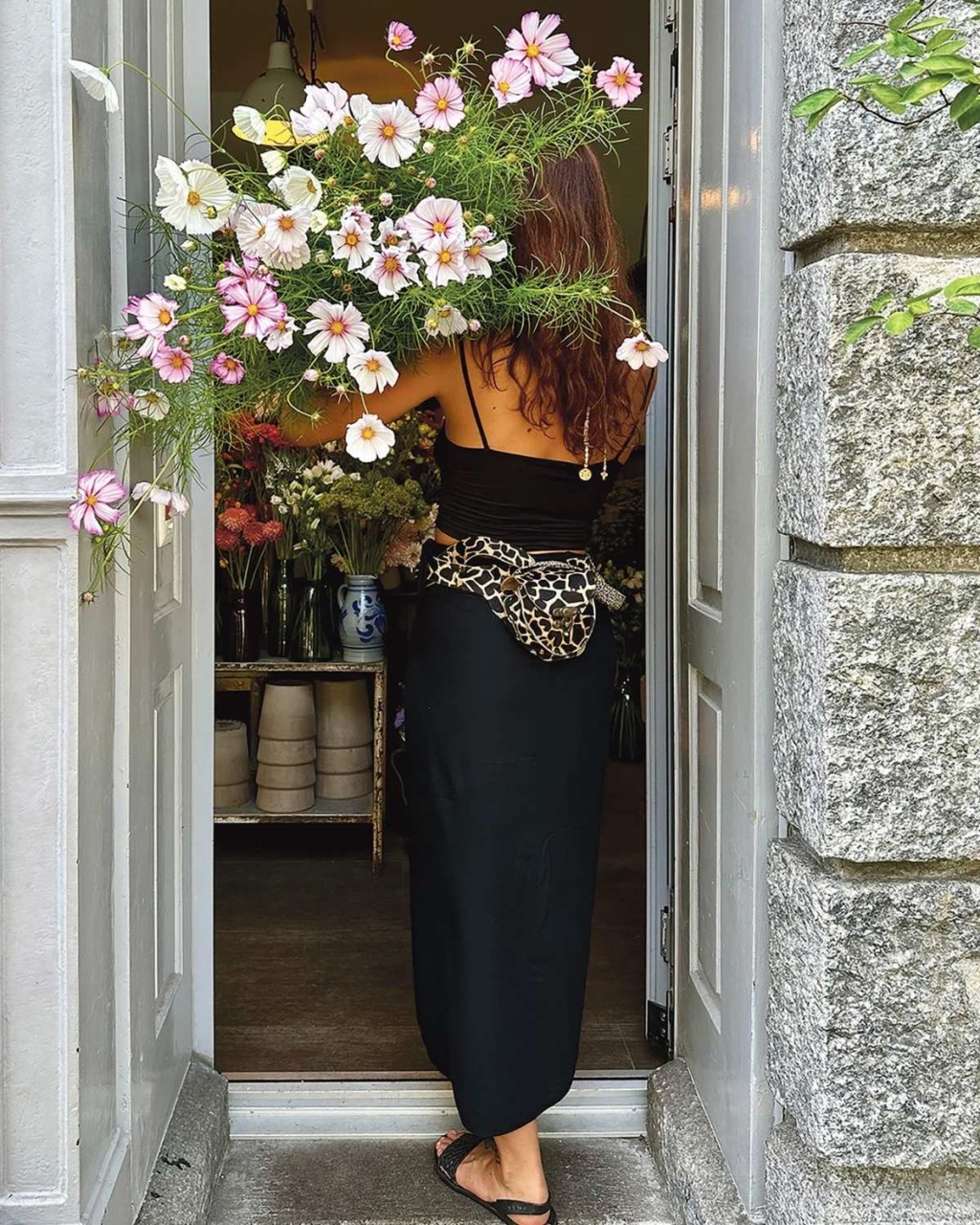 A woman with curly hair stands in a doorway holding a large bouquet of pink and white flowers. She wears a black dress and leopard print waist bag.