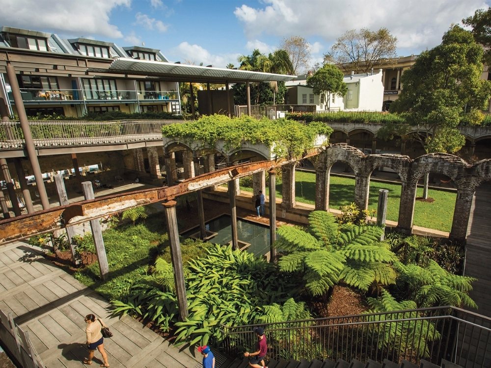 A lush urban park with historic brick arches supports a modern glass building. Greenery, ferns, and walkways create a serene, inviting atmosphere.