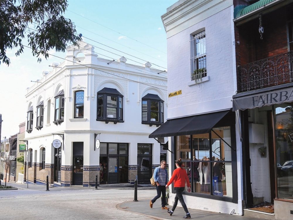 Street corner with historic buildings; a white one with arched windows and a black awning on the right. Two people walking, tree and street sign visible. Bright, relaxed atmosphere.