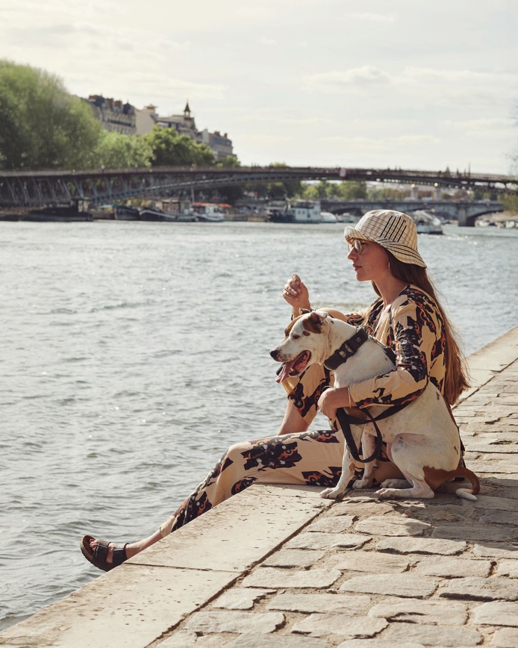 A stylish woman in patterned clothing and a sun hat sits by the riverbank with her dog, gazing at the water. A bridge and cityscape are visible in the background, conveying a peaceful, summery atmosphere.