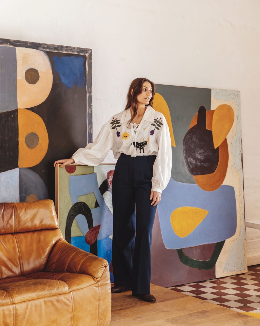 A woman in a white blouse with floral embroidery stands beside abstract paintings in a cozy room with a brown leather chair, exuding a calm, artistic vibe.