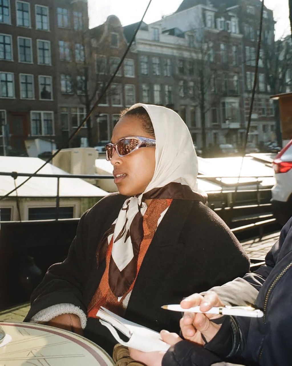 A woman in sunglasses and a headscarf sits at an outdoor cafe table by a canal, with historic buildings in the background. She appears relaxed.