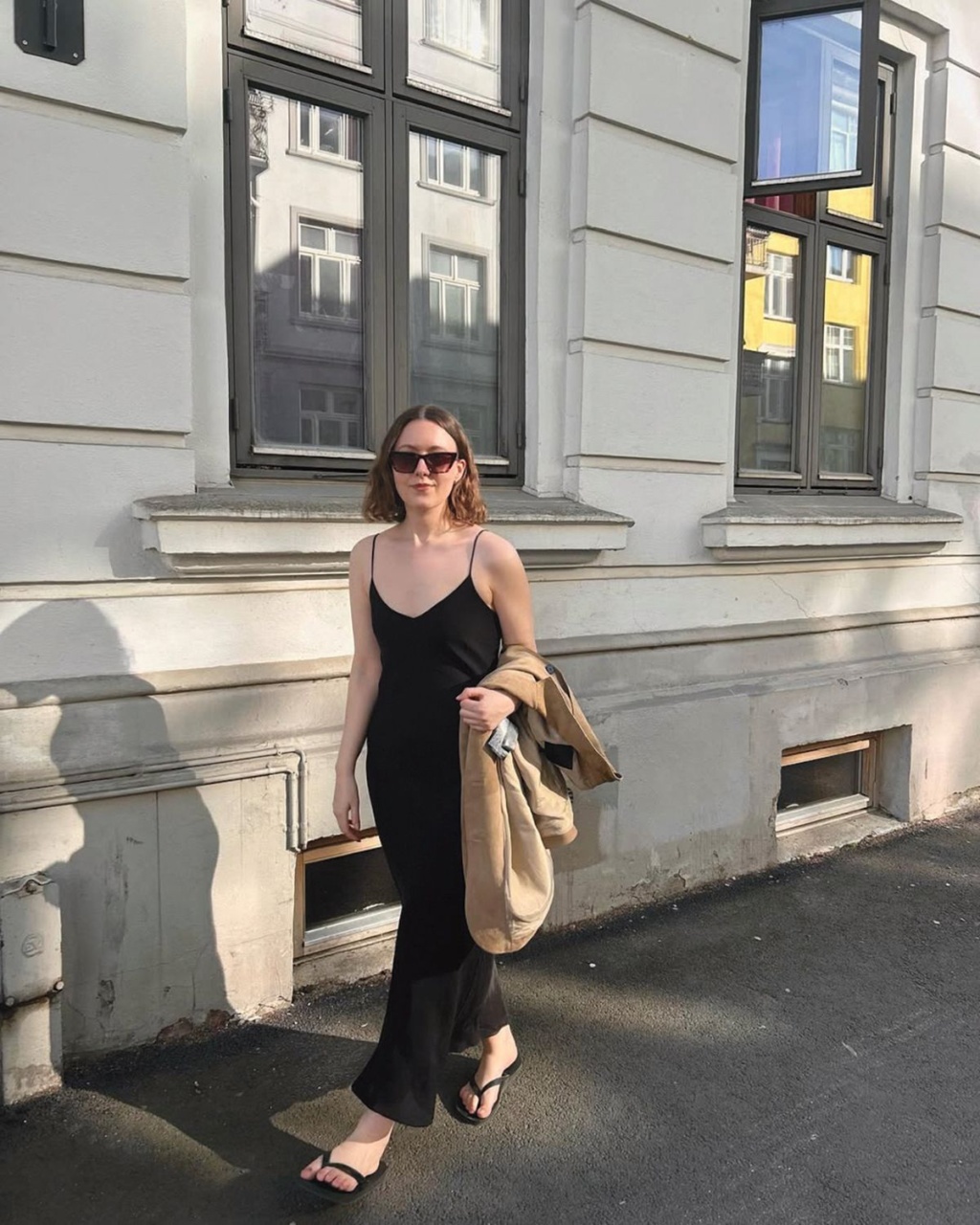 A woman in sunglasses and a black dress walks beside a sunlit building with large windows. She carries a light jacket, exuding a relaxed, stylish vibe.
