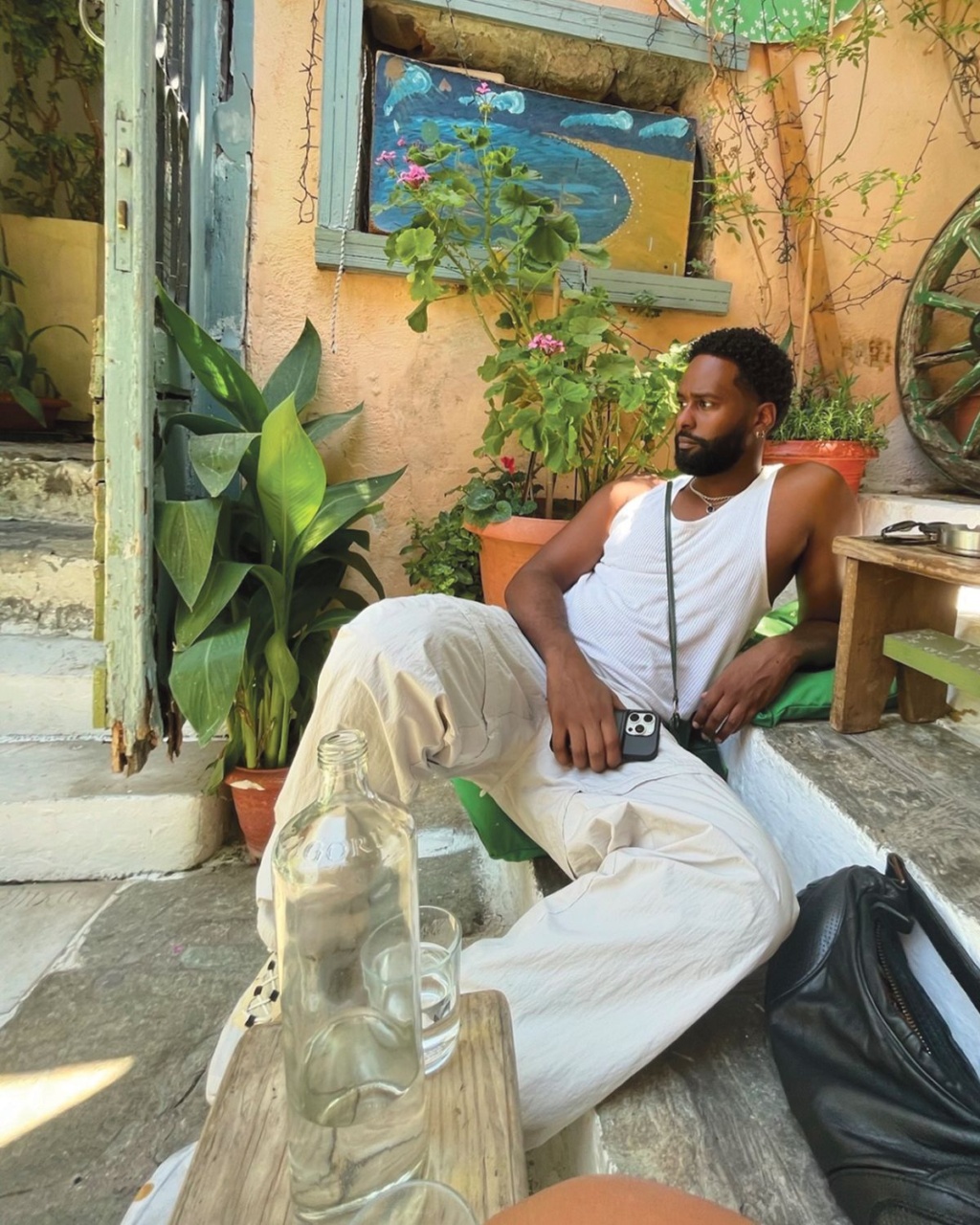 Man in a white tank top and pants relaxes on outdoor steps, surrounded by lush plants. A painted mural and rustic decor create a serene ambiance.