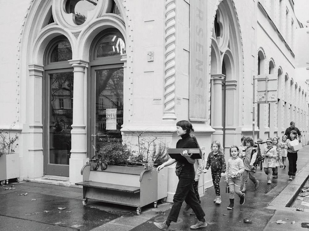 A group of children with an adult walk past a historic building with arched windows and detailed masonry.