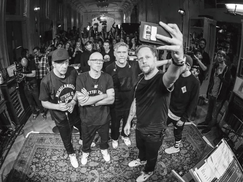 A group of four men stands closely together for a selfie in a room with an ornate ceiling and large crowd behind them.
