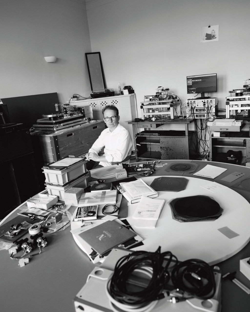 A man in a lab sits at a cluttered table surrounded by vinyl records and tech equipment. The room has an organized yet busy feel.