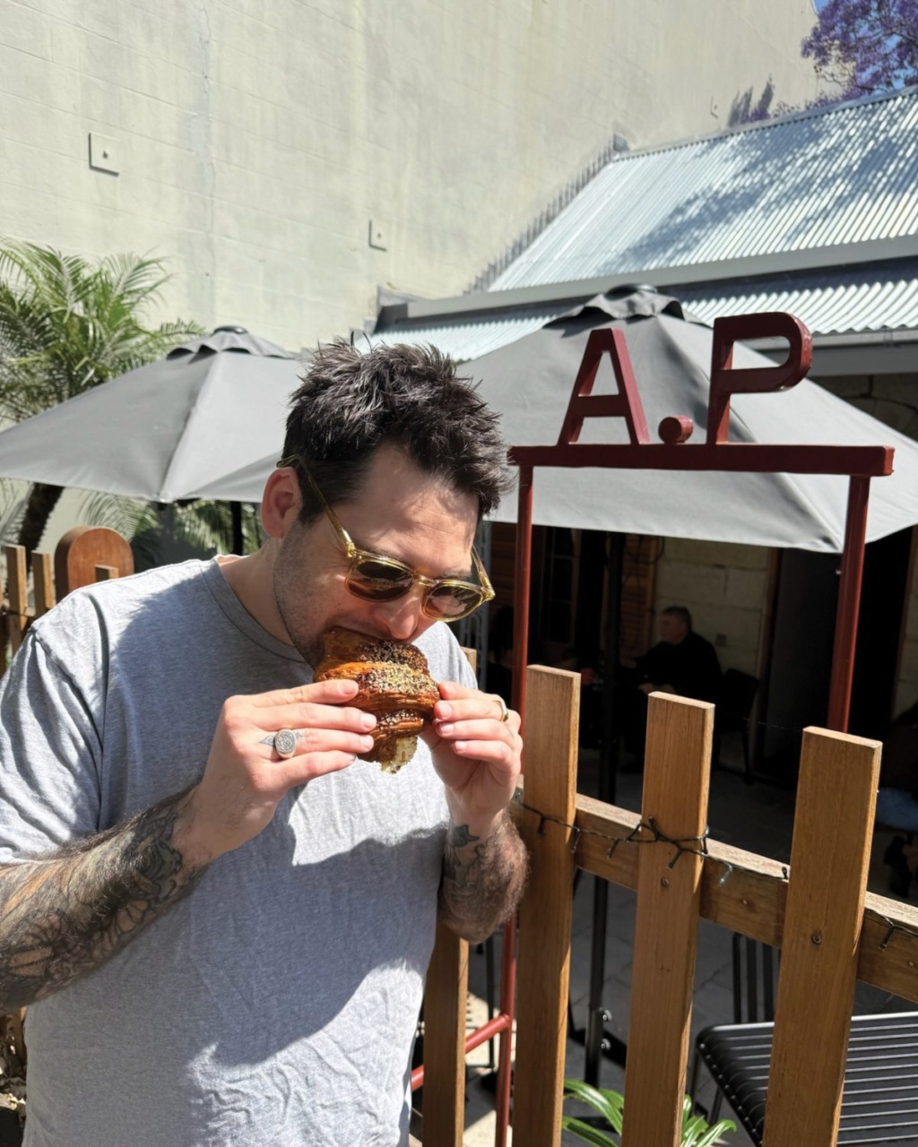 Man enjoying a sesame burger outdoors at a cafe. He wears a gray shirt and sunglasses. Sunlight casts shadows, and a wooden fence is visible.