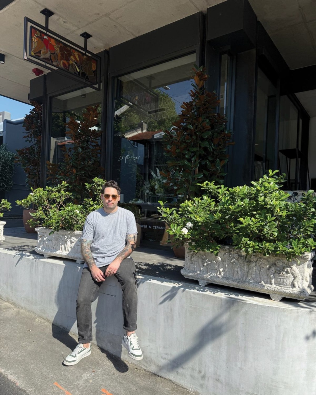 A man with sunglasses sits on a concrete ledge outside a store with large windows. He is surrounded by lush potted plants, giving a relaxed vibe.