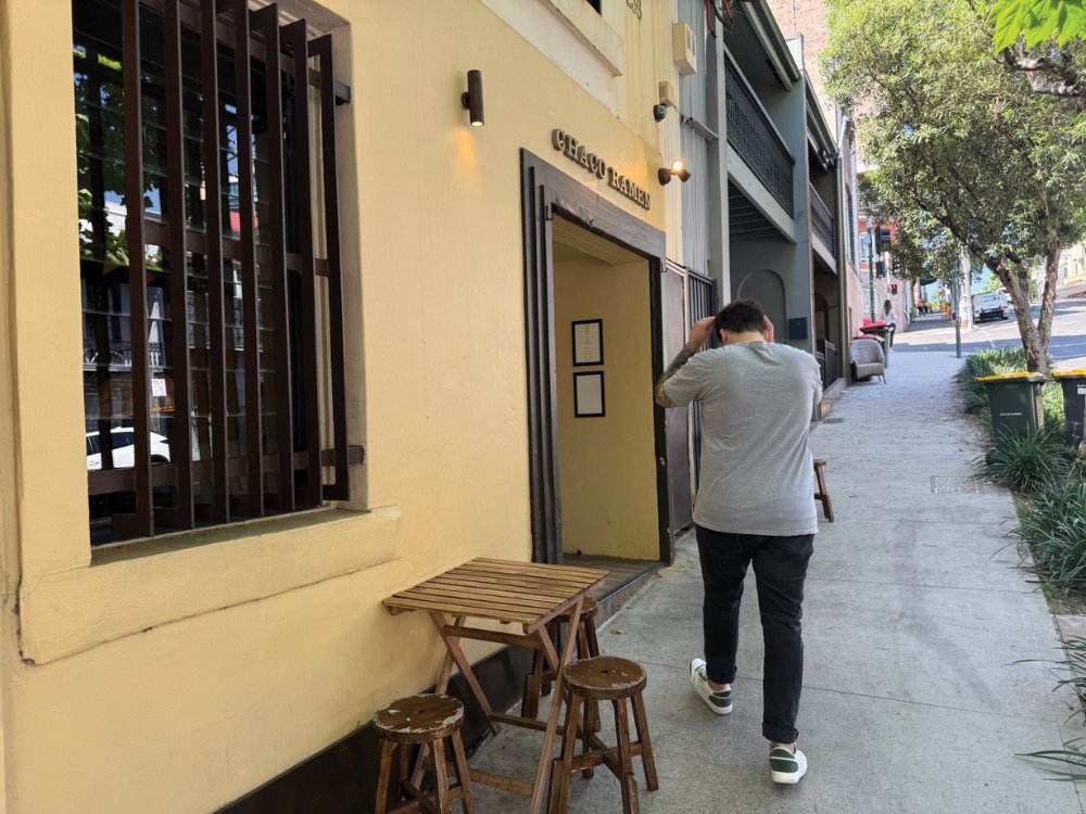 A person in a gray shirt walks past a small cafe with a yellow facade and wooden tables. The street is lined with trees, creating a calm urban scene.