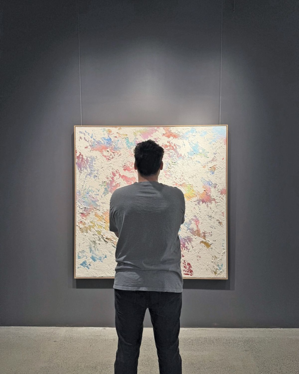 A person stands in front of an abstract painting in a gallery, their back to the viewer. The artwork features colorful, textured patterns on a neutral wall.