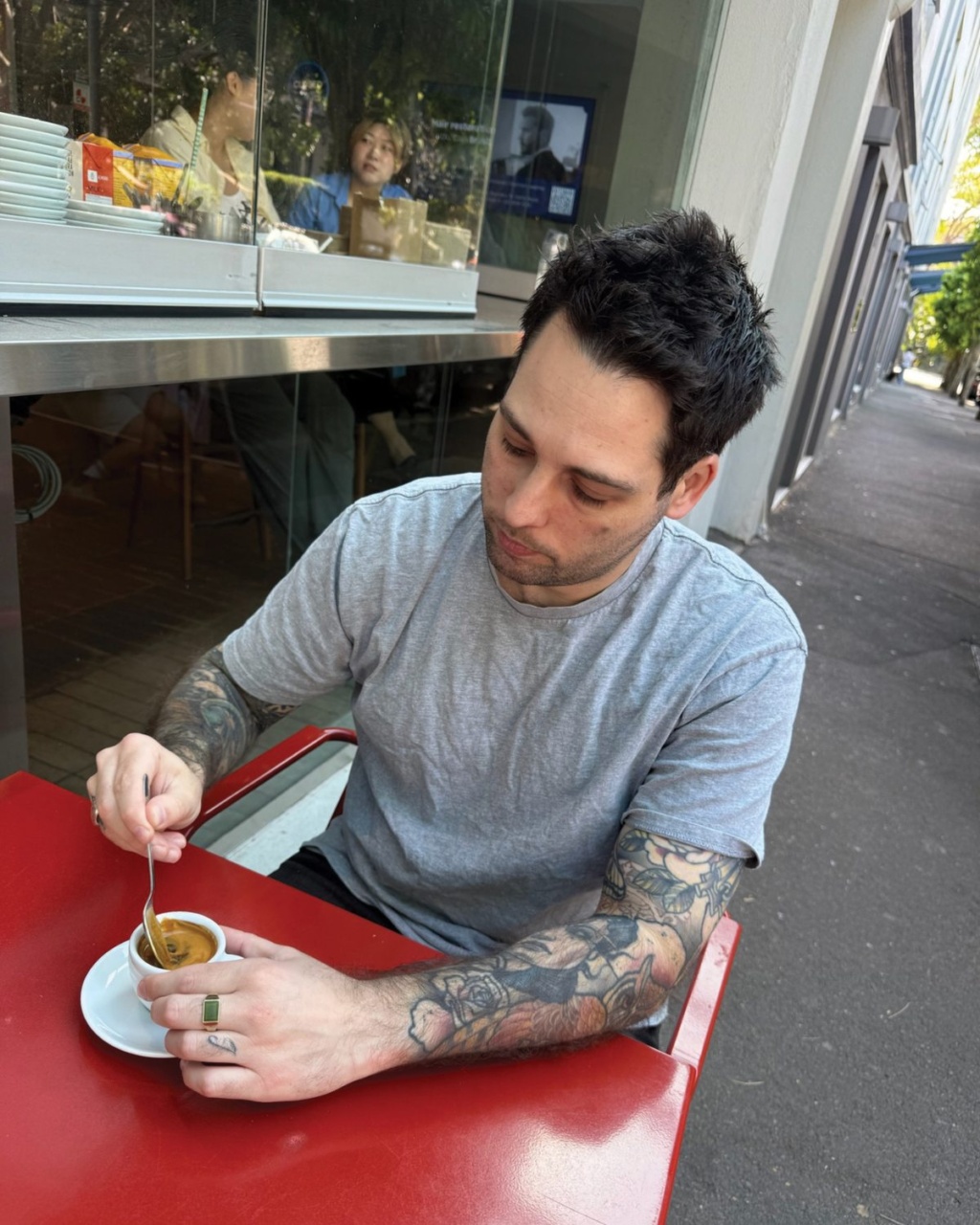 Man with tattoos stirs coffee at a red table outside a café, looking pensive. Reflections of people are visible in the window behind him.