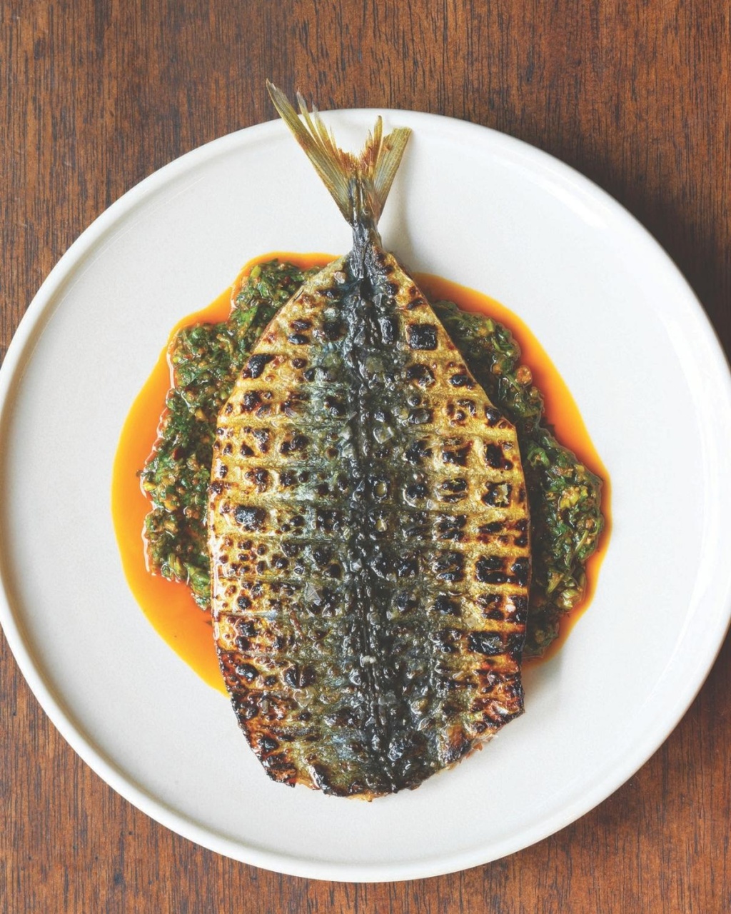 Grilled fish with char marks on a white plate, resting on a bed of vibrant green sauce with a hint of red oil, creating a rustic.