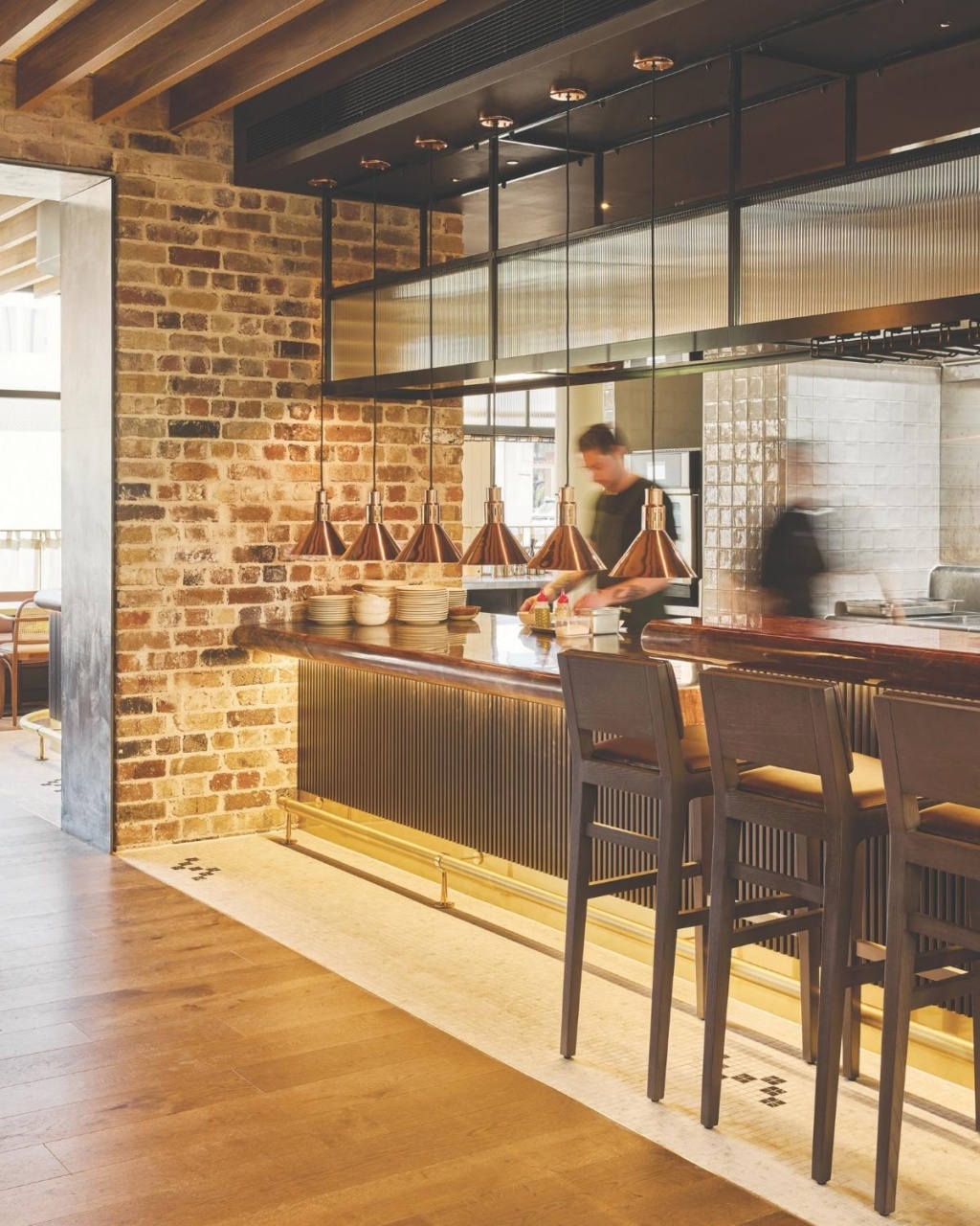 Modern kitchen with brick walls, wooden ceiling, and sleek counter. Copper pendant lights hang above, and a person moves in the background.