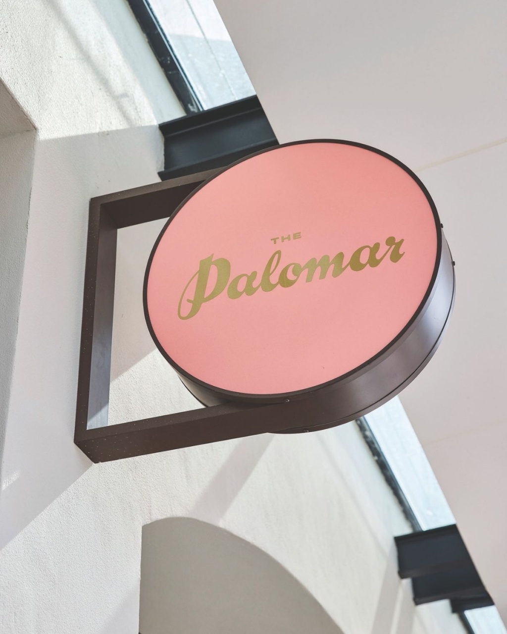 A round, pink sign with "The Palomar" in gold letters hangs on a building's white exterior.