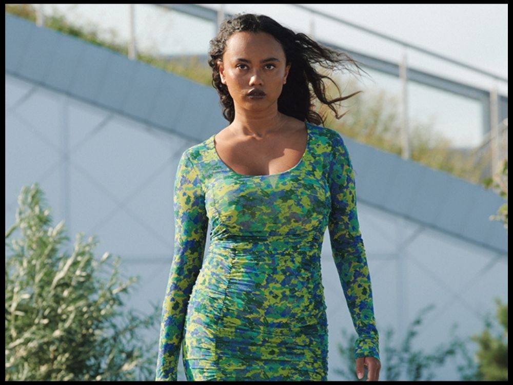 A woman confidently walks outdoors in a fitted, colorful green and blue dress. Her hair is windswept, and she is set against a modern architectural background.