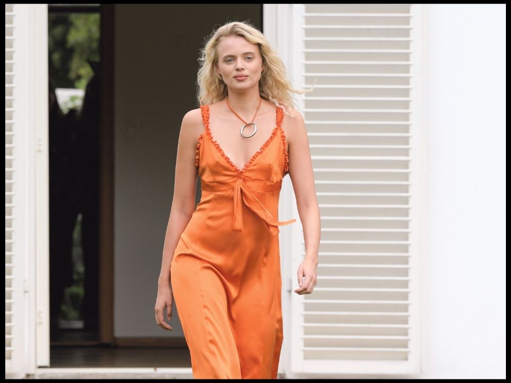 A person with long blonde hair confidently walks outside, wearing an orange dress with a neckline accessory.