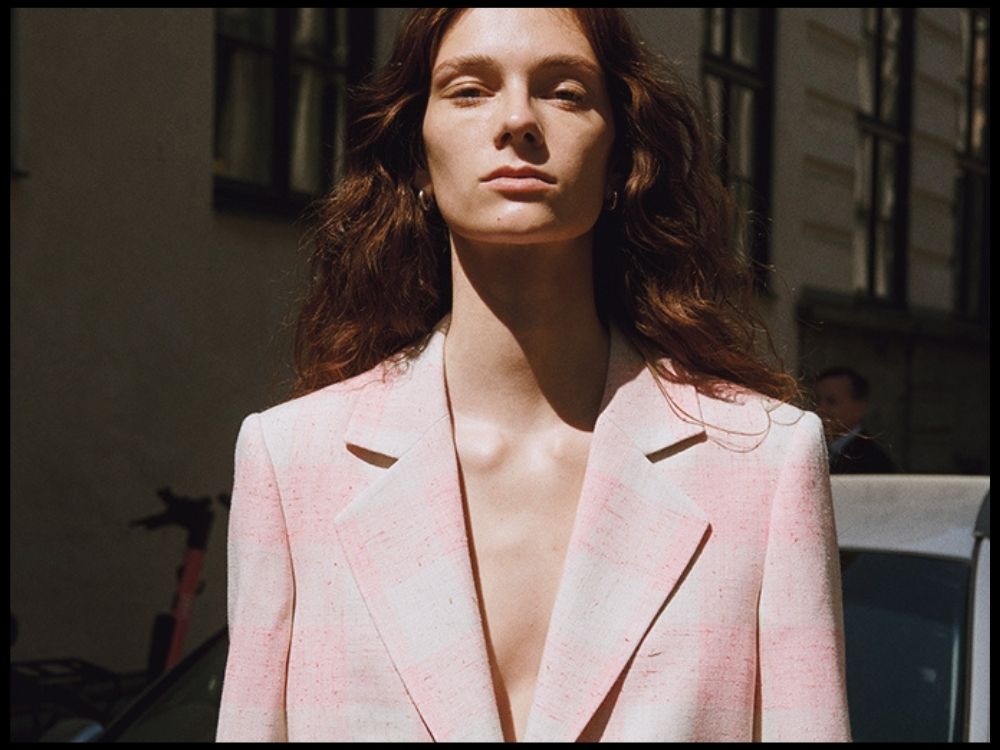 A person with long brown hair stands confidently in sunlight wearing a light pink blazer, with a serious expression.