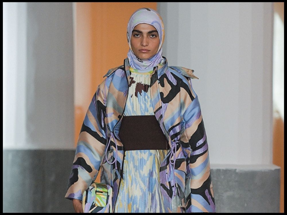 Model in colorful, abstract-patterned coat and hijab walks runway, holding matching bag.