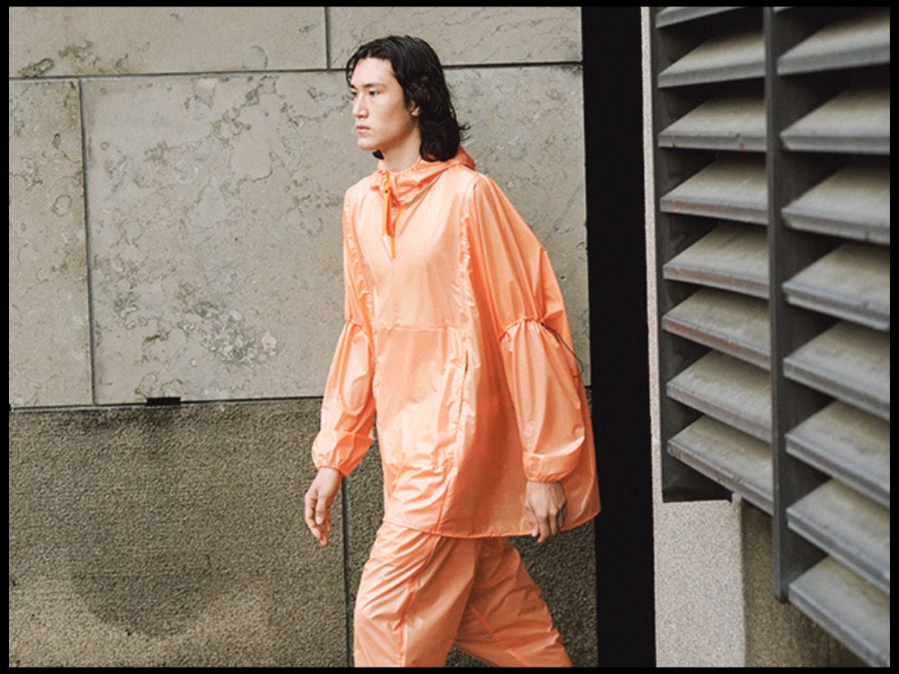 A person with long hair walks in an outdoor urban setting, wearing a shiny, loose-fitting orange tracksuit. The tone is modern and casual.