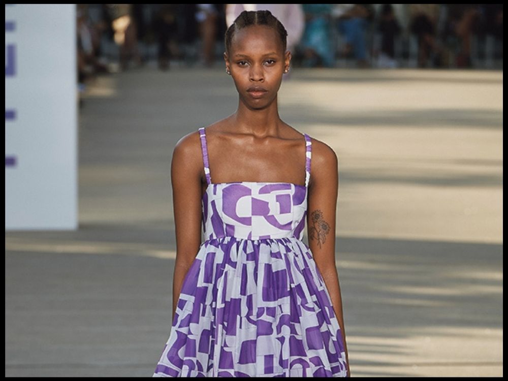 A model wears a black and white striped dress with puffed sleeves and gold buttons, standing outdoors with blurred urban buildings in the background.