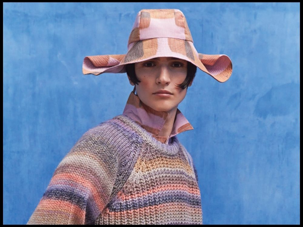 Person wearing a textured, multicolored sweater and a pink plaid hat against a blue background.