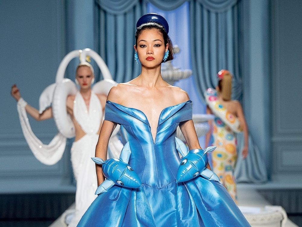 Model in a vibrant blue, off-the-shoulder gown adorned with dolphin-shaped elements, walks a fashion runway; two avant-garde designs are visible in the background.