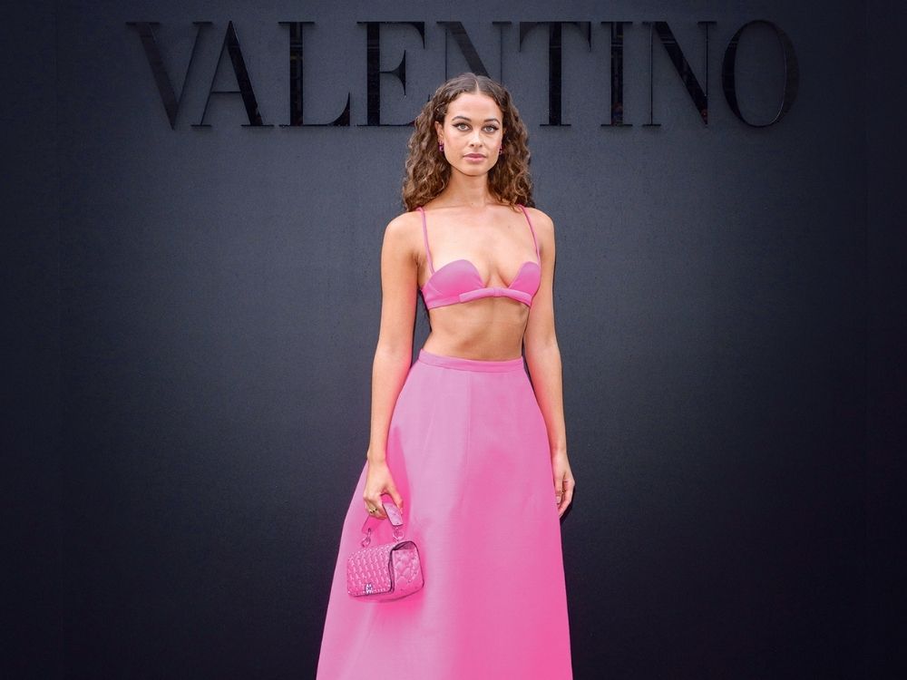 Woman in a pink crop top and skirt holds a matching handbag in front of a black wall with "VALENTINO" text. Elegant and fashionable scene.