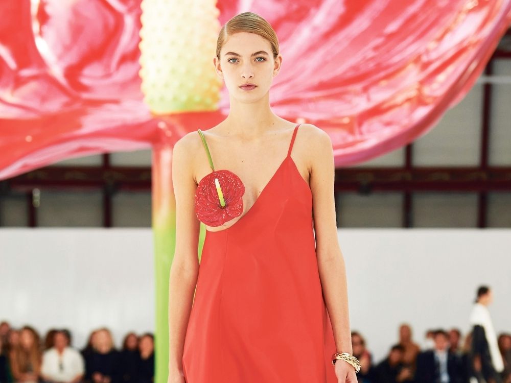 Model in a red dress with a large flower design on the front walks a runway. In the background, oversized red flower petals and a blurred audience create an artistic, vibrant atmosphere.