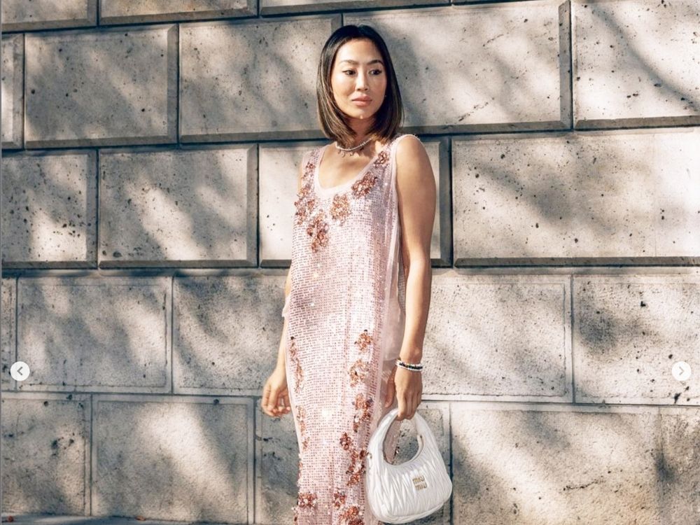 A woman in a shimmering pink sequin dress with floral patterns stands against a textured stone wall. She holds a white bag.