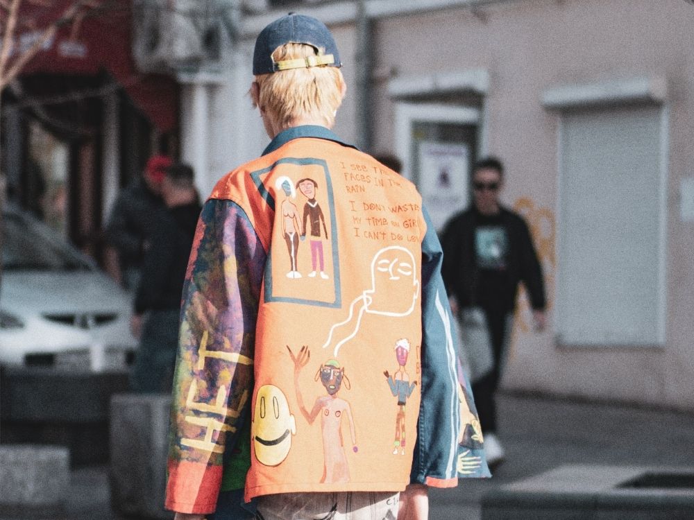 A person with blonde hair wearing a vibrant, graffiti-style jacket featuring abstract faces and text stands on a street, conveying an artistic, urban vibe.
