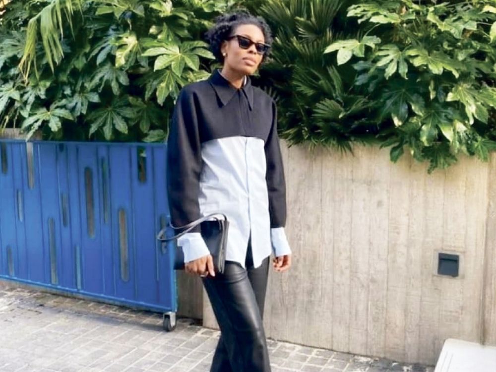 A person wearing a black and white shirt and sunglasses stands casually on a sidewalk, with lush green plants and a blue fence in the background.
