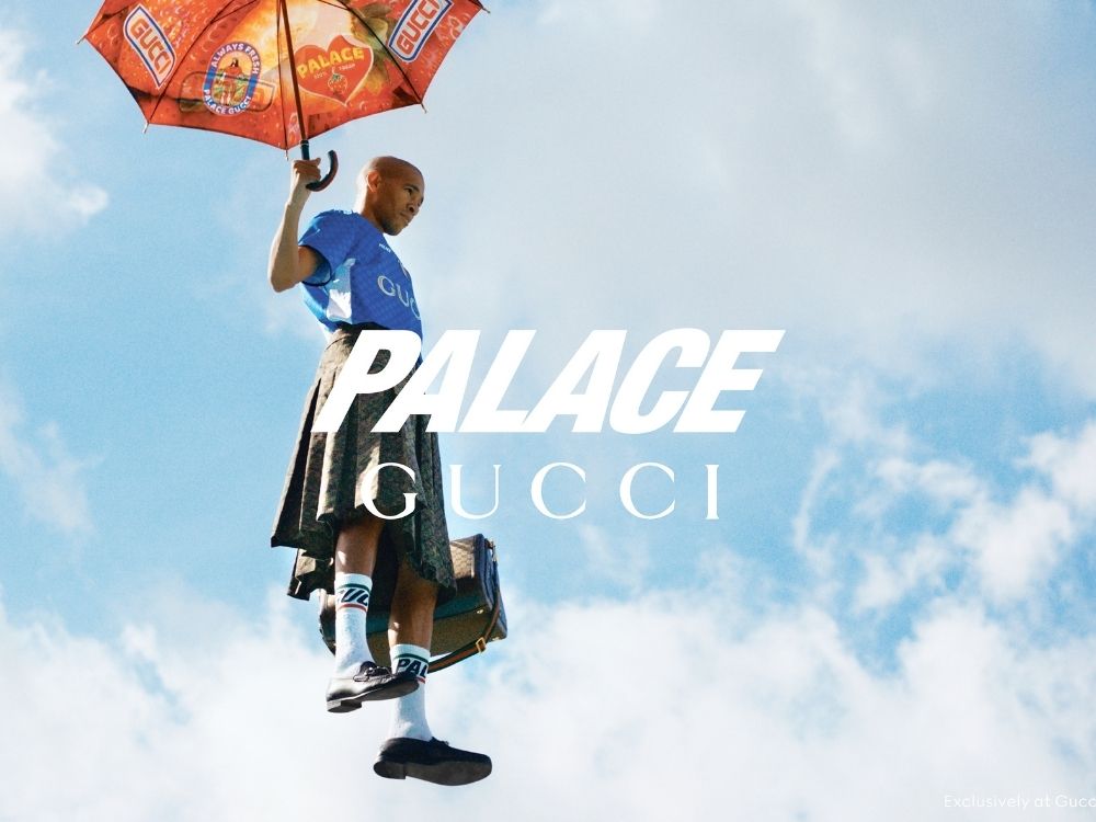 Man in a kilt flies with a Palace Gucci umbrella against a blue sky. He carries a suitcase, wearing socks and shoes, conveying a whimsical tone.